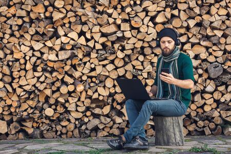 bearded hipster freelancer in headphones working on laptop and talking on phone against background...
