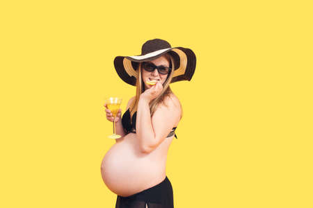 attractive pregnant woman on a yellow background holds a glass of martini in her hands and bites a piece of lemon.の写真素材