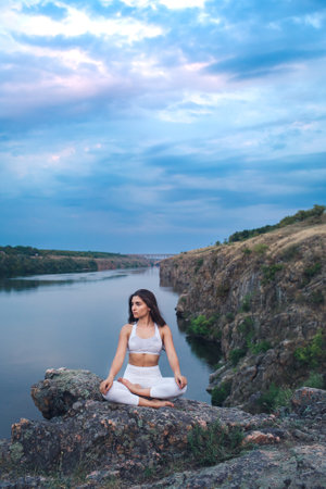 a young girl sits in a yoga lotus meditation position on the river bank on a rockの写真素材