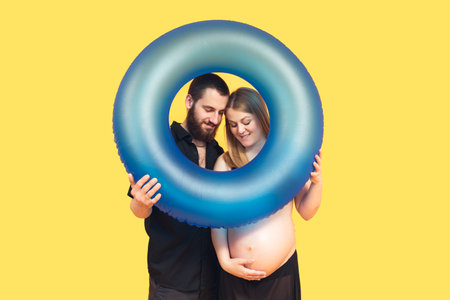 Portrait of a cheerful positive pregnant couple with a blue swim ring, enjoy their vacation on a bright yellow backgroundの写真素材