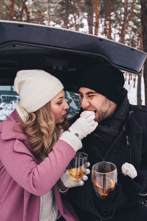 Picnic in the winter in the car. A couple drinks tea from a . The girl feeds the guy marshmallows.の写真素材