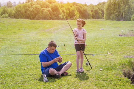 dad and his daughter are preparing gear for fishing, near the pond in the meadow, the girl looks and learnsの写真素材