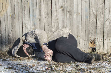 Homeless. At the wooden fence lies the body of a man in dirty and torn clothes with a medical mask on his face. close up.の写真素材