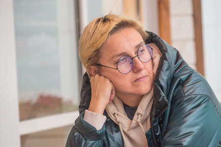 a girl with short hair and glasses is sitting in a cafe leaning on a table thinking about something portrait in warm colors.の写真素材