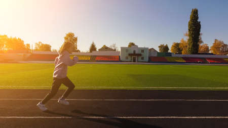 a little teenage girl runs fast on the treadmill of the stadium near the football field.の写真素材