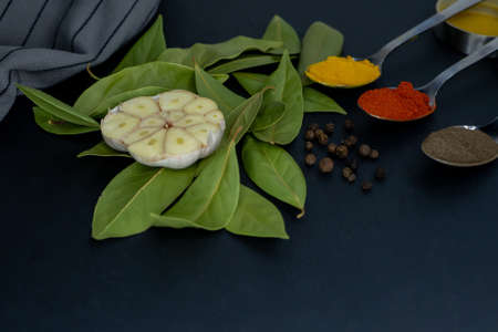 Side view half a garlic bulb on bay leaves, pepper peas, spoons with seasonings and a towel on a dark background. Close-up.の写真素材