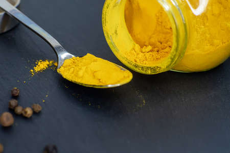 Jar and spoon with turmeric and peppercorns on a dark background, side view. Close-up.