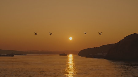 Birds fly gracefully across an orange-tinted sky at sunset over a calm body of water with distant silhouettes of hills.の素材