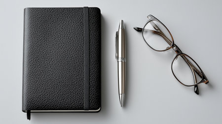 A neatly arranged set of a black leather-textured notebook, a sleek silver pen, and a pair of clear-lens glasses on a smooth gray surface. The composition, captured from a top-down perspective, showcases minimalist elegance, with soft natural lighting highlighting the textures and reflective surfaces.の素材