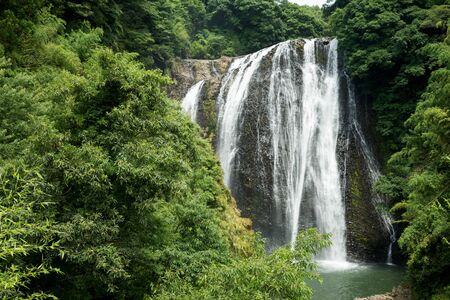Waterfall falling along the bedrock in back of green forestの写真素材