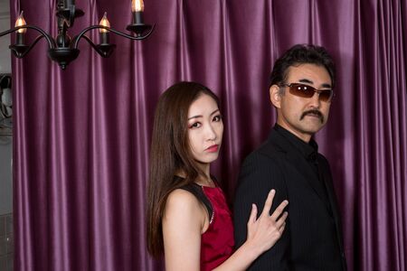 Red dressed woman snuggling up to suited man in front of purple curtainの写真素材