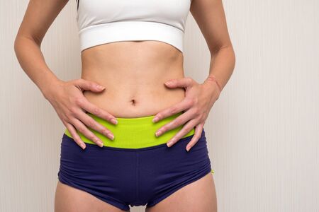 Close up abdomen of sportswear woman her hands on side of the navel in front of light gray wallの写真素材