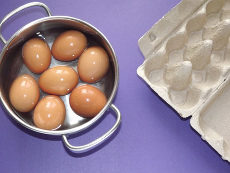 beige boiled eggs in a pot of water with an egg box on a purple background. High quality photo