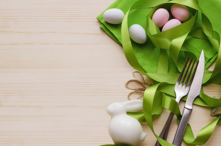 White decorative rabbit, colorful eggs, line napkin and cutlery with green ribbon on wooden background with copy space. Top view. Happy Easter concept.の写真素材