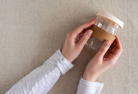 Female hand holding reusable glass coffee cup on natural linen background. Reuse, reduce, recycle...