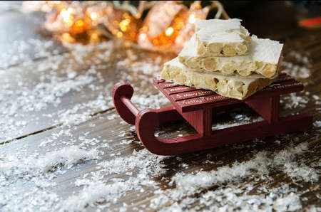 Turron, traditional Spanish sweet consumed at Christmas. Almond nougat dessert on decorative sleigh with snow and Christmas tree with garland on the background.Copy space.の写真素材