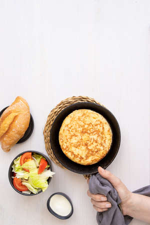 Women hand holding frying pan with Tortilla, Spanish omelette made with eggs and potatoes and served with salad and bread. Top view.の写真素材