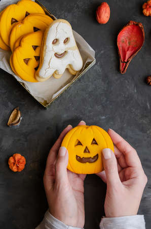 Female hands holding pumpkin scary face cookie on dark background with halloween symbol sand decoration. Happy Halloween concept.の写真素材
