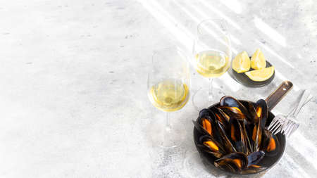 Steamed mussels served in frying pan, glasses of white wine on cement background. Popular...