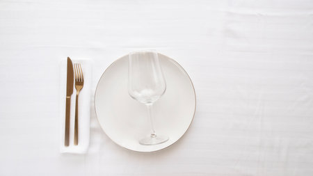 Gold cutlery knife and fork in porcelain plate and wine glass on white tablecloth for restaurant...