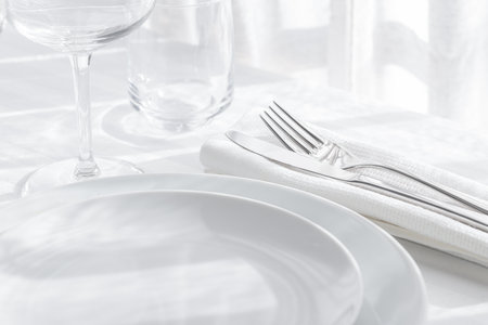 Table setting white and gray colour. Empty glasses and plates set with napkin and cutlery on white table cloth. Restaurant interior background.の写真素材