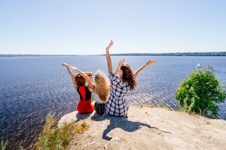 Shoot of three beautiful girls outdoors by the river. Female friends relaxing by the river and smiling. Girlfriends are stand on the pier, joy, fun, hands up. Girls with drinks in dresses. Summerの写真素材