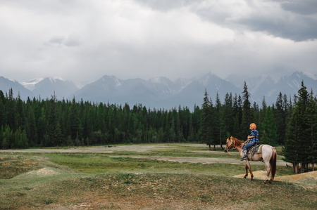 young girl with red head riding by horse. Beautiful green forest and big mountains on the background.の写真素材