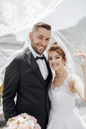 elegant beautiful couple under veil. Hugs on each other. They look into the camera and smile sincerely. Love in the eyes. Gorgeous light adds a romantic atmosphereの写真素材