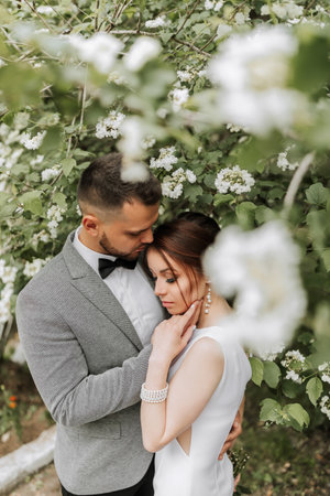 the bride in a white evening dress poses with the groom with a beautiful hairstyle, the groom gently wraps the bride and leans against her against the background of spring flowers.の写真素材