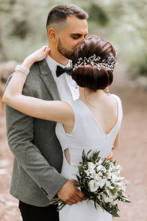 The groom holds a bouquet of flowers and hugs his wife, kissing her temple. The bride has her back turned. Photo detailsの写真素材