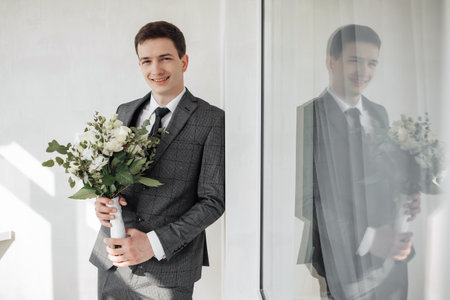 The groom, standing, holding a delicate wedding bouquet, smiling sincerely. Beautiful sunlight. Wedding portrait.の写真素材