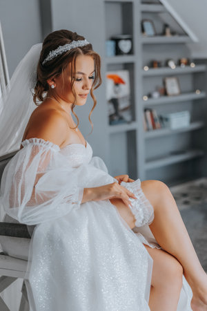 Young bride in a white dress with a wedding garter on her leg, close-up. Preparation for the wedding celebration. Accessories for the newlyweds, lace garter.の写真素材