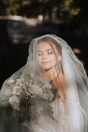 Beautiful curly brown-haired bride in a white dress poses for a photographer, standing under a veil in a beautiful dress with sleeves. Wedding photography, close-up portrait, chic hairstyle.の写真素材