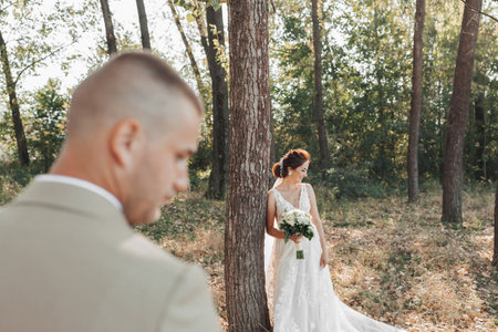 Wedding photo in nature. The bride is standing near a tree in a beautiful dress, holding a bouquet of white roses, looking down. The bridegroom is in the foregroundの写真素材
