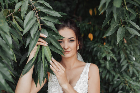 Wedding photo on a green background. The bride in a white dress stands near green bushes, holds a leaf near her face and looks into the lens. Beautiful makeup. Portrait of the brideの写真素材