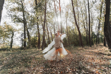 Portrait of happy bride and groom in the forest. The groom holds the bride in his arms, dancing with her. A long veil in the air. A ray of the sun in the photo. Wide angle photoの写真素材
