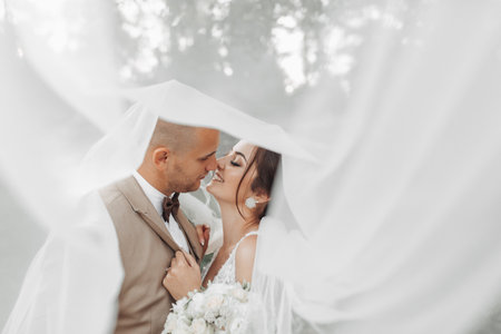 Wedding portrait of the bride and groom. Happy bride and groom gently hug each other under the veil, pose and kiss. Stylish groom. Beautiful young brideの写真素材