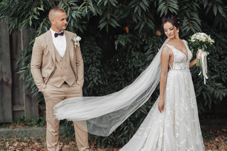 Wedding photo on a green background. The bride and groom are standing near green bushes. Stylish groom in full height. A beautiful bride with a long veil that is thrown in the air. Wedding portraitの写真素材
