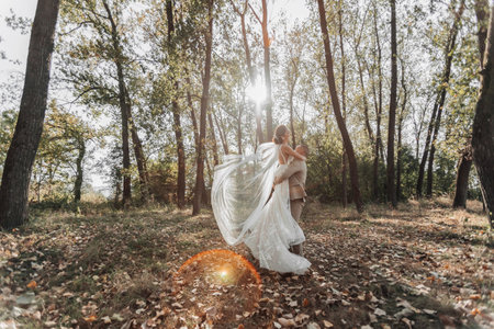 Portrait of happy bride and groom in the forest. The groom holds the bride in his arms, dancing with her. A long veil in the air. A ray of the sun in the photo. Wide angle photoの写真素材
