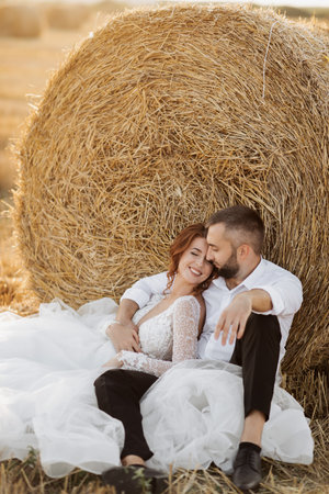 Wedding portrait. The bride and groom are sitting in an embrace on the ground, near a bale of hay. Red-haired bride in a long dress. Stylish groom. Summer. A sincere smile. On the background of hayの写真素材