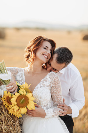 Wedding portrait of the bride and groom. The groom, tearing his shirt, stands behind the bride, near a bale of hay. Red-haired bride in a long dress with a bouquet of sunflowers. Stylish groom. Summerの写真素材