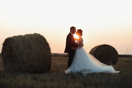 Wedding portrait of the bride and groom. Bride and groom stand embracing at sunset near hay bales. Red-haired bride in a long dress. Stylish, bearded groom. Silhouette photo.の写真素材