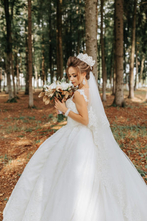 A beautiful young woman in a wedding dress between tall trees in the forest with a royal hairstyle and a chic tiara with a bouquet of flowers in her hands, a wedding in golden colorの写真素材