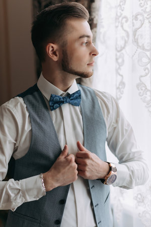 A man in a white shirt and a gray vest poses by the window and looks to the side, adjusts his vest. Front view. A stylish watch. Men's style. Fashion. Businessの写真素材