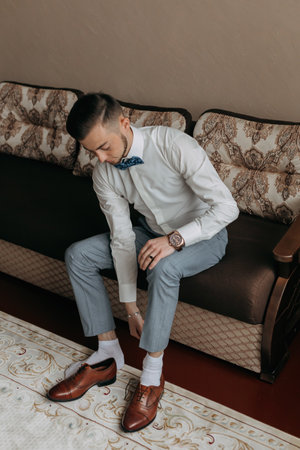 portrait. A man in a white shirt and gray pants is sitting on the sofa and putting on his elegant shoes. A stylish watch. Men's style. Fashion. Businessの写真素材