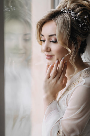 Blonde bride with elegant hairdo and tiara, wearing a robe, posing, looking at the camera, touching her face. Beautiful makeup. Elegant hairstyle. French manicureの写真素材