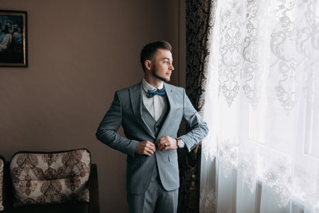 A man in a white shirt and a gray suit poses by the window and looks away. adjusts his jacket. Front view. A stylish watch. Men's style. Fashion. Businessの写真素材
