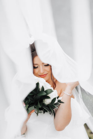 Curly brunette bride in a white dress, covered with a veil, poses for the camera with a bouquet of roses. Portrait of the bride. Beautiful makeup and hair. Wedding in natureの写真素材