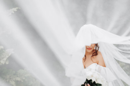 Curly brunette bride in a white dress, covered with a veil, poses for the camera with a bouquet of roses. Portrait of the bride. Beautiful makeup and hair. Wedding in natureの写真素材