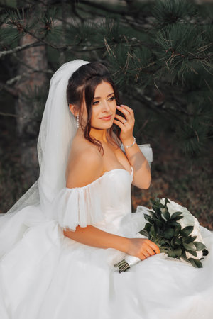 portrait of the bride in nature. A brunette bride in a white voluminous dress is sitting, posing near a coniferous tree, holding a bouquet of white roses. Beautiful hair and makeup. Warm lightの写真素材
